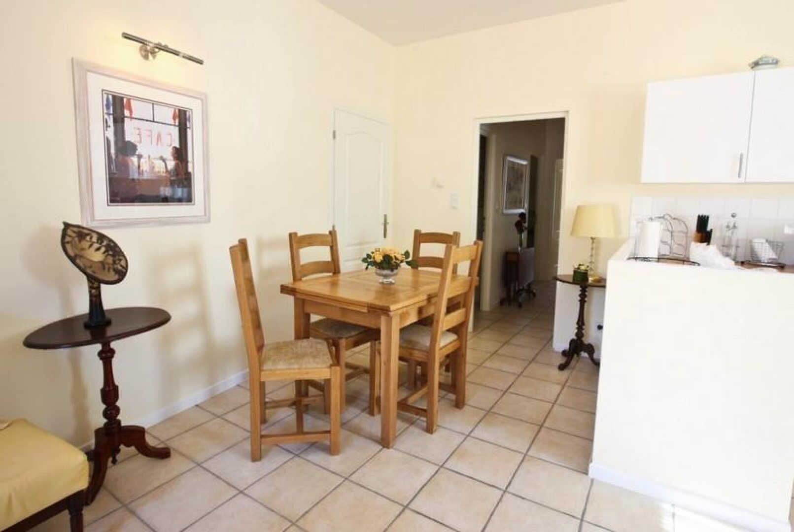 Un coin repas avec une table et des chaises en bois, une petite table ronde agrémentée d’un objet décoratif, et un espace cuisine avec des placards et un réfrigérateur.