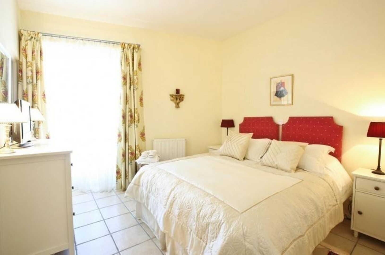 A hotel room with a large bed, a bedside table with a lamp, a radiator, and a window with curtains.