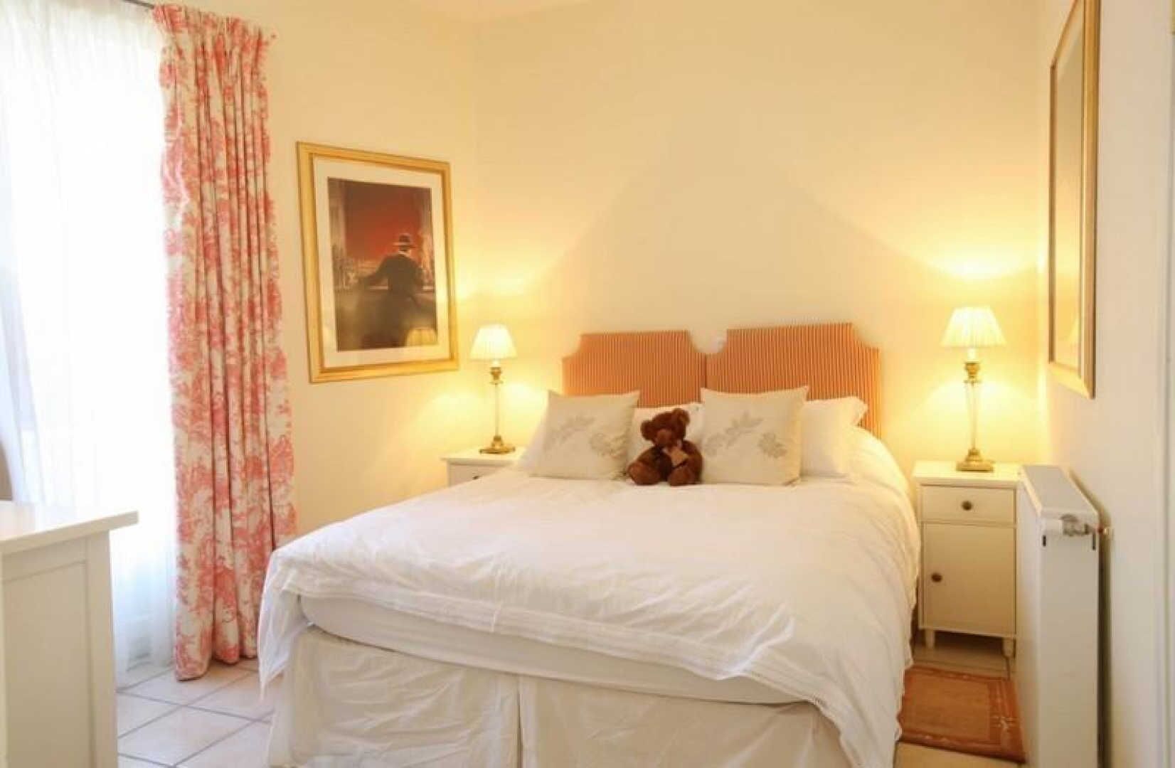 Una cama bien hecha con sábanas blancas, dos lámparas de noche, un osito de peluche sobre las almohadas, un cuadro en la pared y una ventana con cortina estampada.