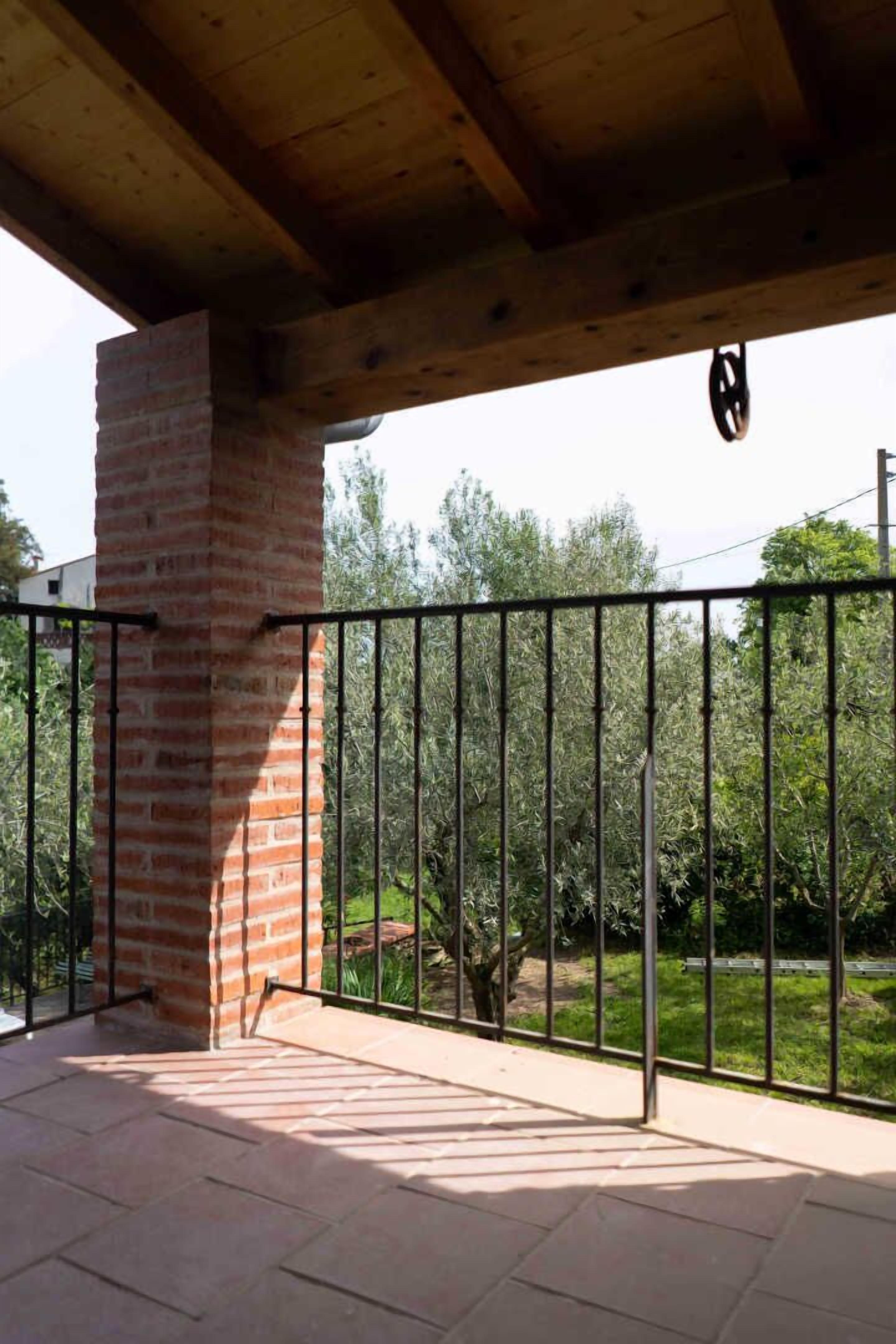 A covered patio area with a brick chimney, a metal railing, and a view of a garden with trees and a fence.