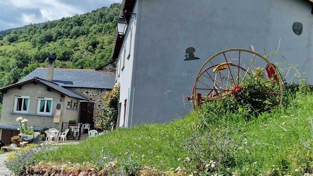 Un bâtiment rustique, doté d’une grande roue à eau fixée sur le côté, entouré d’une végétation luxuriante et adossé à un paysage montagneux.