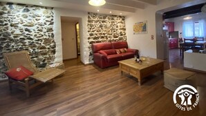 A cozy living room with a stone wall, a red sofa, wooden furniture, and a kitchen in the background.