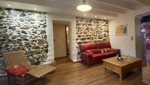 A cozy living room with a stone wall, a red sofa, wooden furniture, and a kitchen in the background.