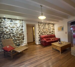 Un salon chaleureux avec un mur en pierre, un canapé rouge, des meubles en bois et une cuisine en arrière-plan.
