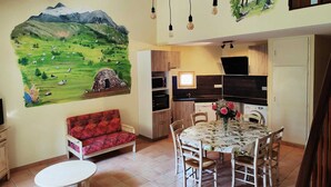 Een gezellige woonkamer met een bank, een eettafel met stoelen, een keukenhoek en een muurschildering van een groen landschap.
