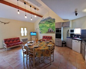 Uma área de refeições com mesa posta para quatro pessoas, um sofá, uma TV e uma cozinha com máquina de lavar.