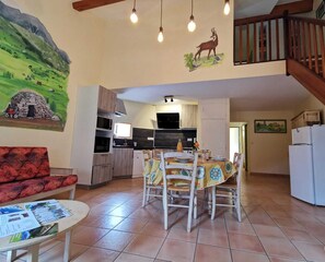 Uma sala de estar aconchegante com um sofá vermelho, uma mesa de jantar posta para quatro pessoas e uma cozinha ao fundo.