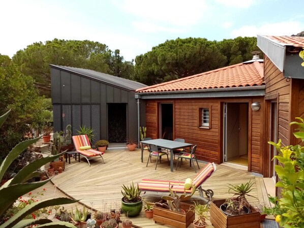 A wooden house with a tiled roof, a deck with lounge chairs, and potted plants.