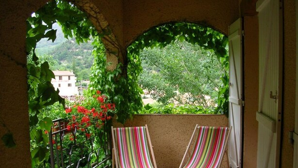 Un balcon avec deux fauteuils de jardin à rayures, une vue sur la verdure et les bâtiments, et un mur en pierre.