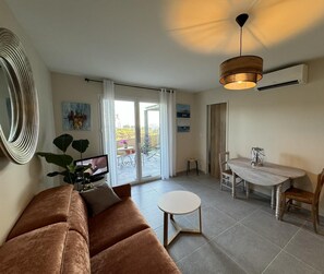 A living room with a sofa, a small round table, and a dining table with chairs. There is a sliding glass door leading to an outdoor area with a balcony.