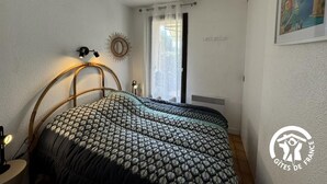 A bedroom with a bed, a nightstand, a wall-mounted lamp, a window with curtains, and a painting on the wall. - Gîte de la Lagune - SAINT CYPRIEN (SAINT CYPRIEN)
