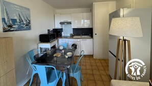A compact kitchen with white cabinets, a microwave, a coffee maker, and a washing machine. There is a dining table with blue chairs and a painting of sailboats on the wall. - Gîte de la Lagune - SAINT CYPRIEN (SAINT CYPRIEN)
