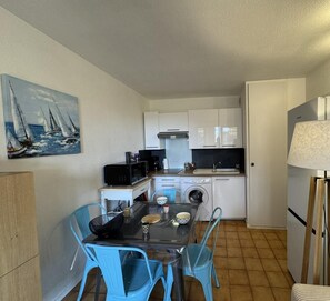 A compact kitchen with white cabinets, a microwave, a coffee maker, and a washing machine. There is a dining table with blue chairs and a painting of sailboats on the wall.