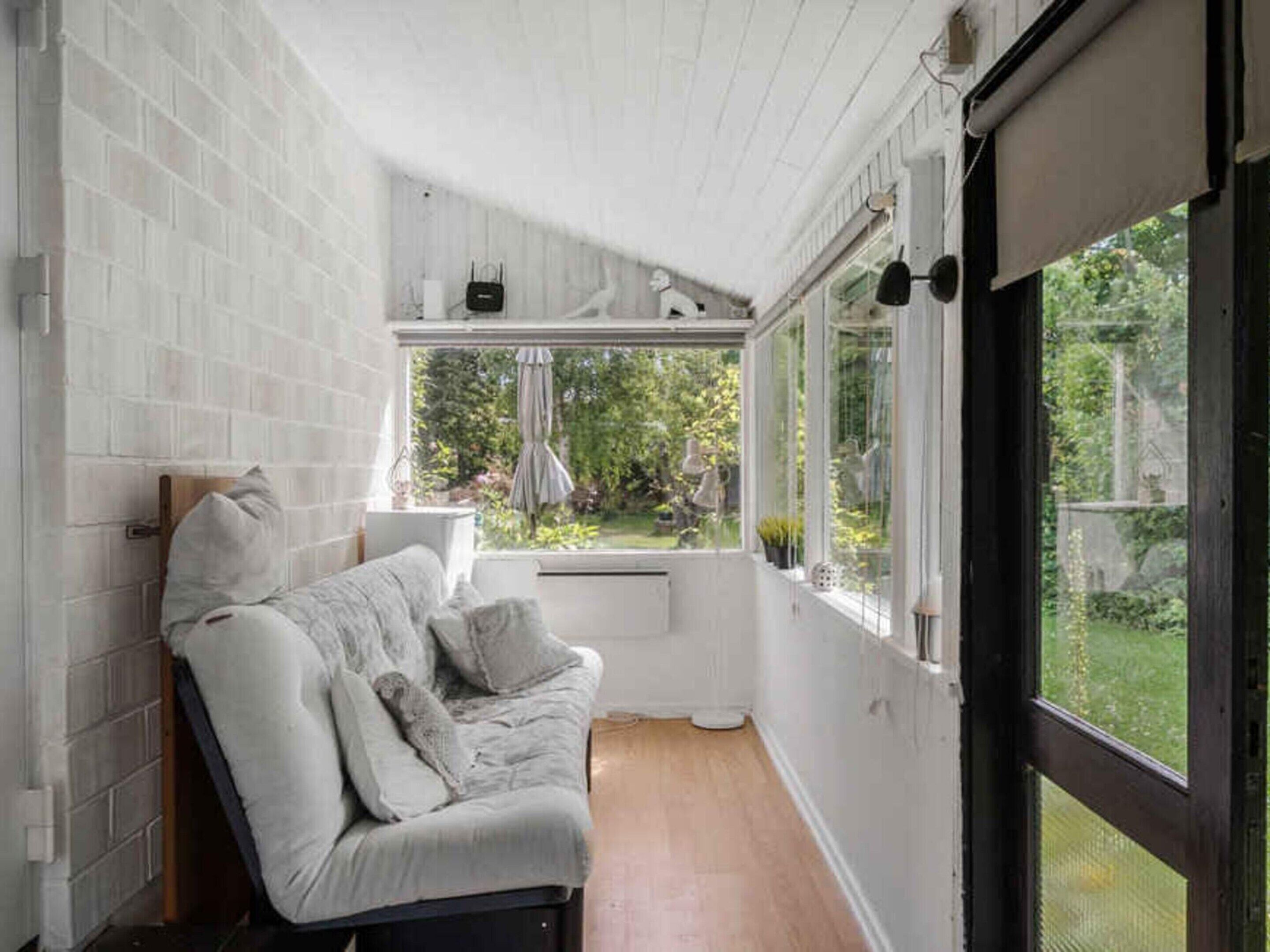 A cozy room with a white brick wall, a wooden floor, a white sofa with cushions, a window with a view of greenery, and a small shelf with decorative items.