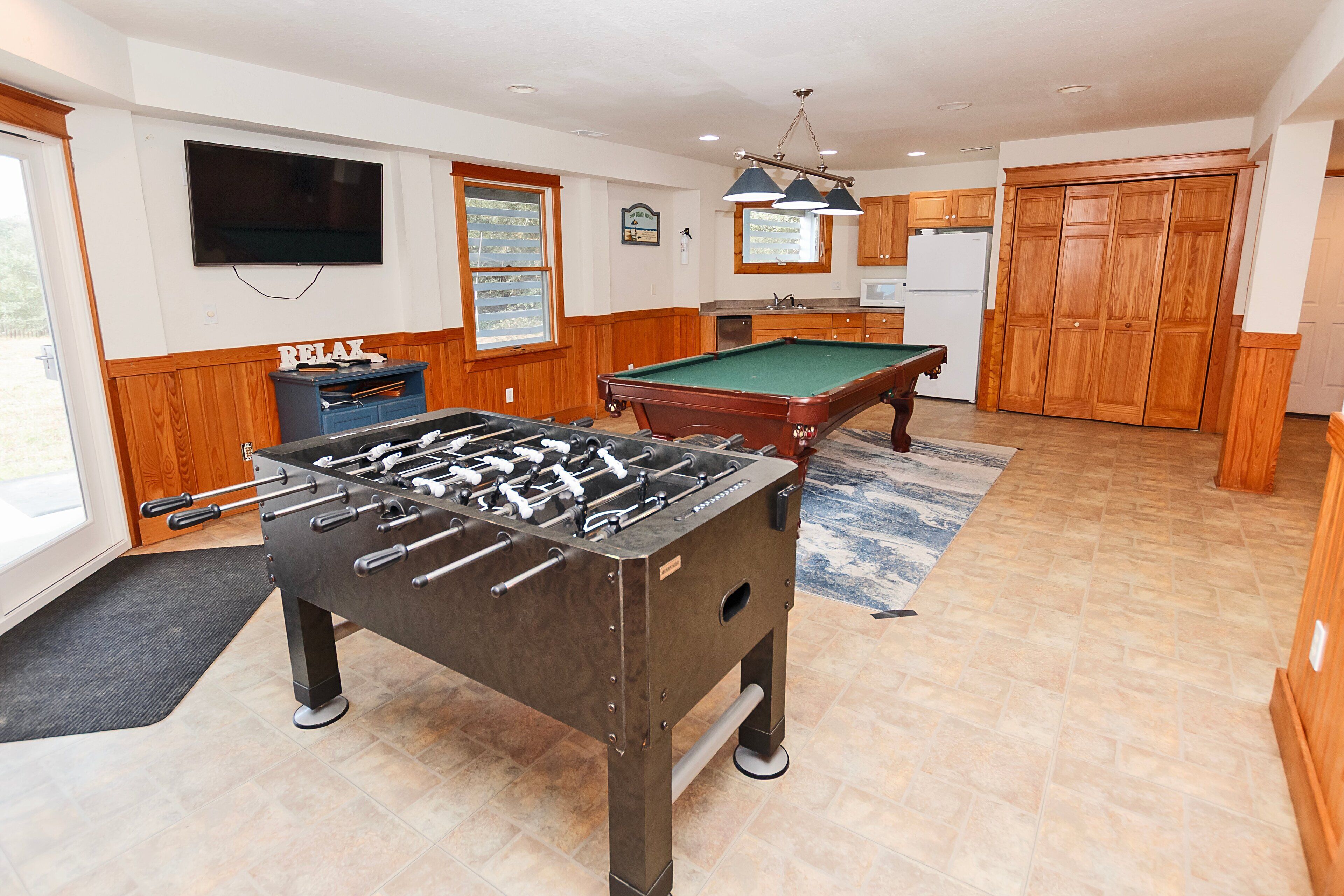 A room with a foosball table, a pool table, wooden cabinets, a television, and a kitchen area.