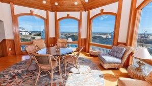 Amplia habitación con un gran comedor rodeado de sillas de mimbre, un sofá acolchado y un tapiz estampado. Amplias ventanas en arco ofrecen vistas panorámicas del paisaje.