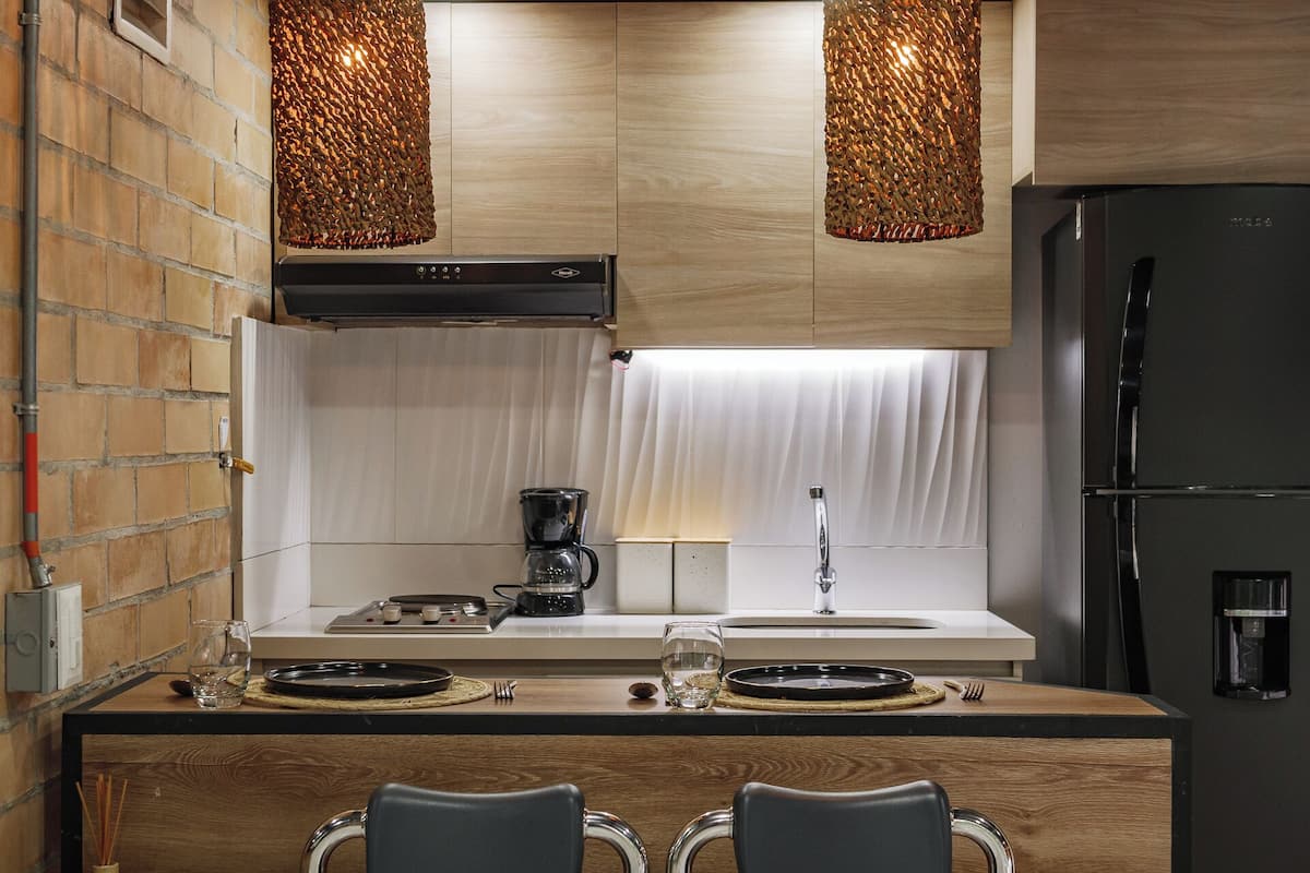 A compact kitchen with a brick wall, wooden cabinets, a black refrigerator, and two pendant lights.