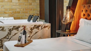 Quarto de hotel com toucador revestido de mármore, cama com roupa de cama branca, garrafa de vinho sobre um tabuleiro de madeira e uma luminária moderna.