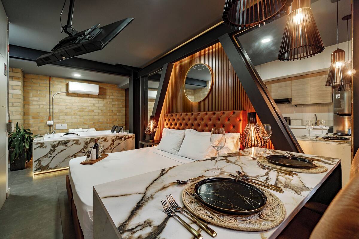 A modern hotel room with a large bed, a separate bathroom, a dining area with a marble table, and a kitchen in the background.