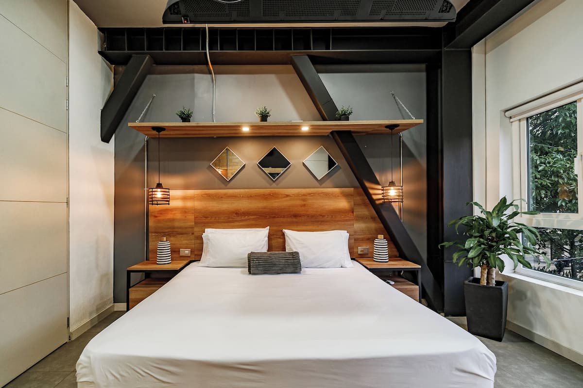 A modern bedroom with a large bed, wooden headboard, bedside lamps, and a view of greenery through a window.