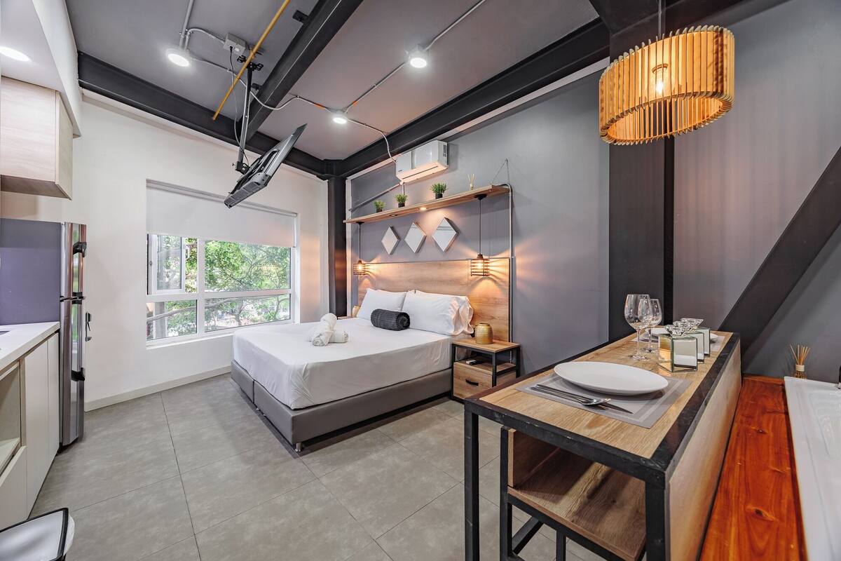 A modern hotel room with a bed, bedside table, and a dining area.
