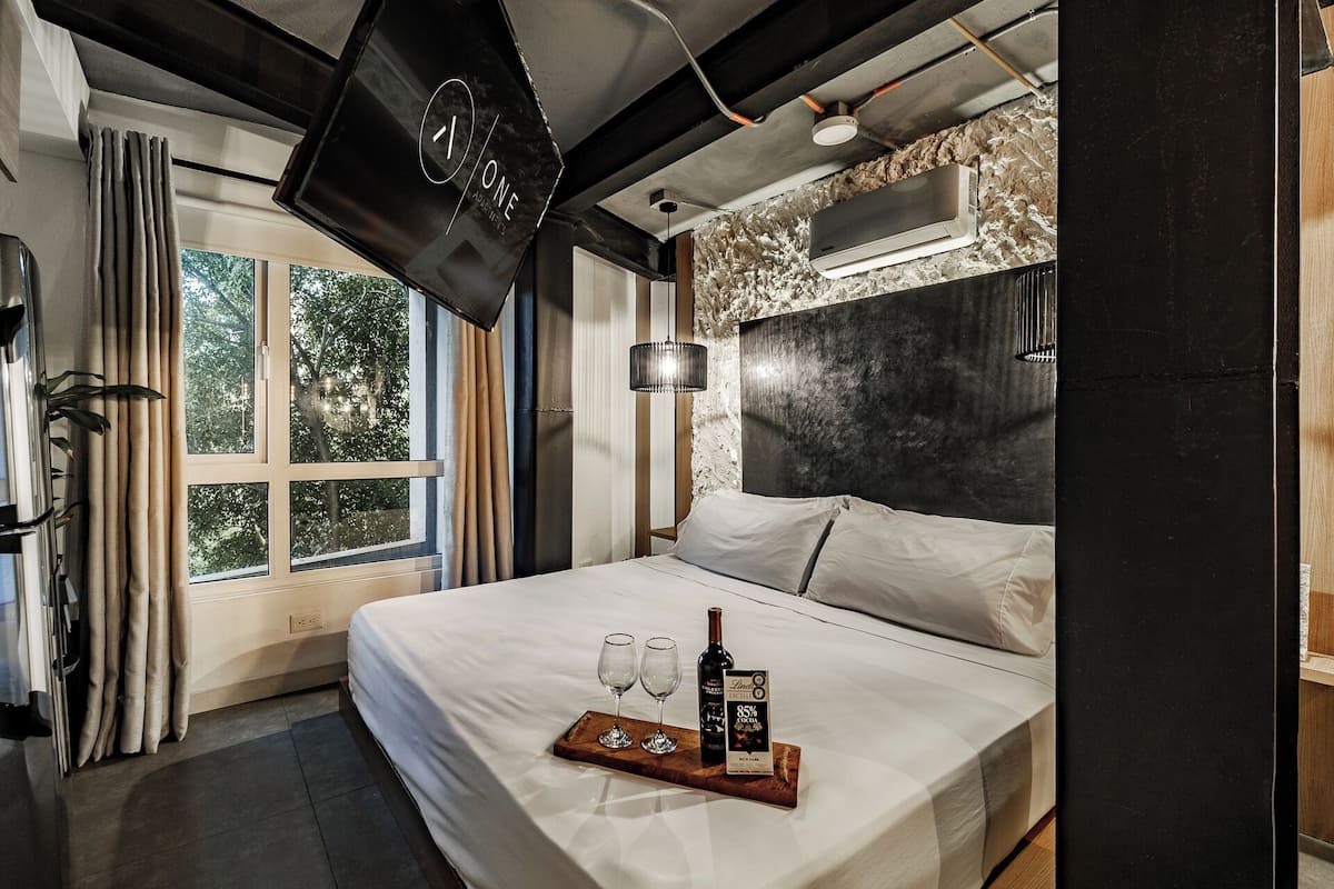 A modern hotel room with a large bed, a mounted TV, a window with curtains, and a tray with a bottle and glasses.