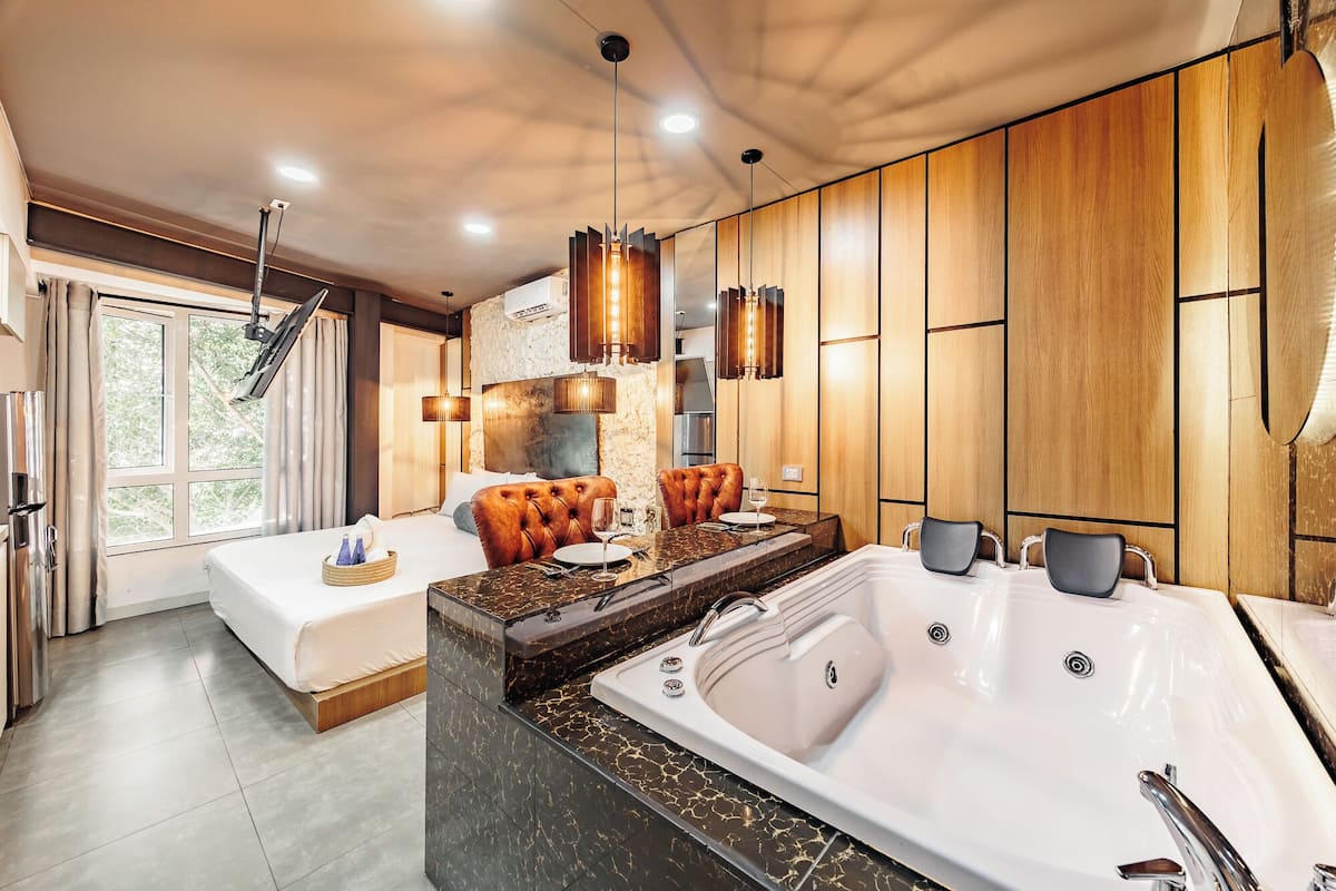 A modern hotel room with a large bed, a sitting area with a sofa and table, a separate bathroom with a bathtub, and a view of greenery outside.