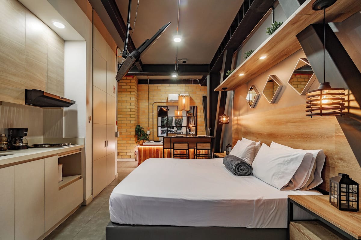 A modern bedroom with a large bed, bedside tables, a desk, and a kitchen area in the background.