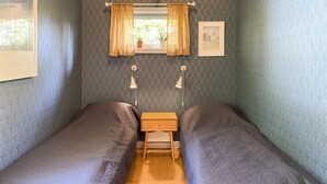 A small, cozy room with two single beds, a nightstand, a window with curtains, a wall-mounted lamp, and a framed picture.