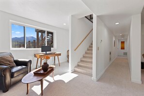 Uma sala de estar moderna com sofá, mesa de centro, escrivaninha com computador e vista para montanhas através de grandes janelas.