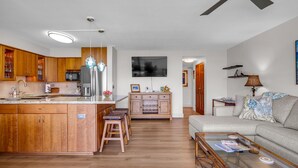 Et moderne køkken med træskabe, en central ø med barstole, et spiseområde med et glasbord og en stue med sofa og lampe.