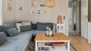 A cozy living room with a grey sectional sofa, a wooden coffee table, and a patterned rug. There are framed pictures on the wall and a wicker lamp on a side table. - 5 Star Holiday Home in Dronningmolle (Dronningmolle)