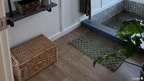 Un baño con suelo de madera, una alfombrilla de baño verde, un estante con velas y un florero, una bata colgada en un perchero y una ventana con vistas.