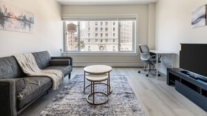 A modern living room with a grey sofa, a round table, a TV, and a large window with a city view.