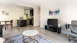 A modern living room with a marble coffee table, a TV on a black stand, a dining area with a wooden table and chairs, and a kitchen in the background.