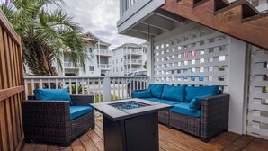 A wooden deck with wicker furniture and a table, overlooking a white building with balconies.