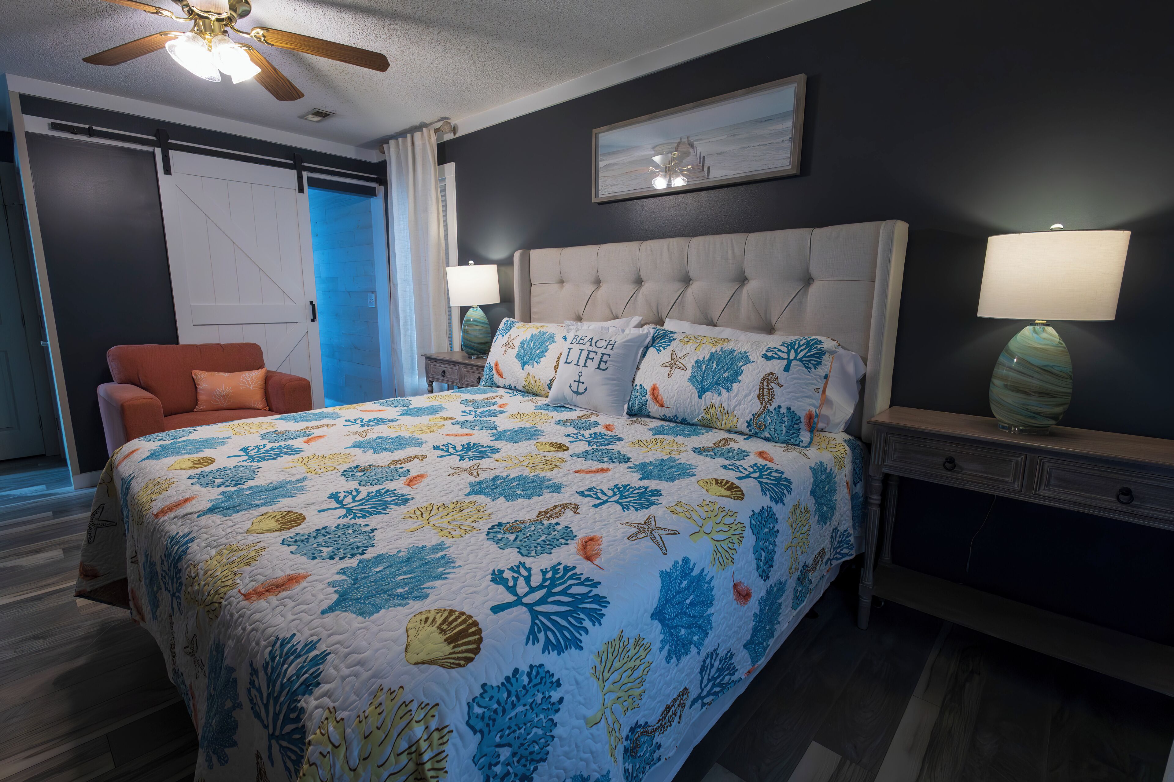 A bedroom with a bed, a chair, a nightstand, a lamp, and a ceiling fan.