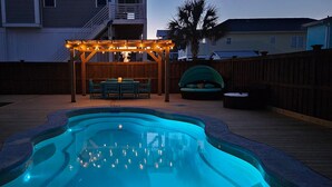 A swimming pool with a wooden deck, a covered seating area with string lights, and a palm tree in the background.