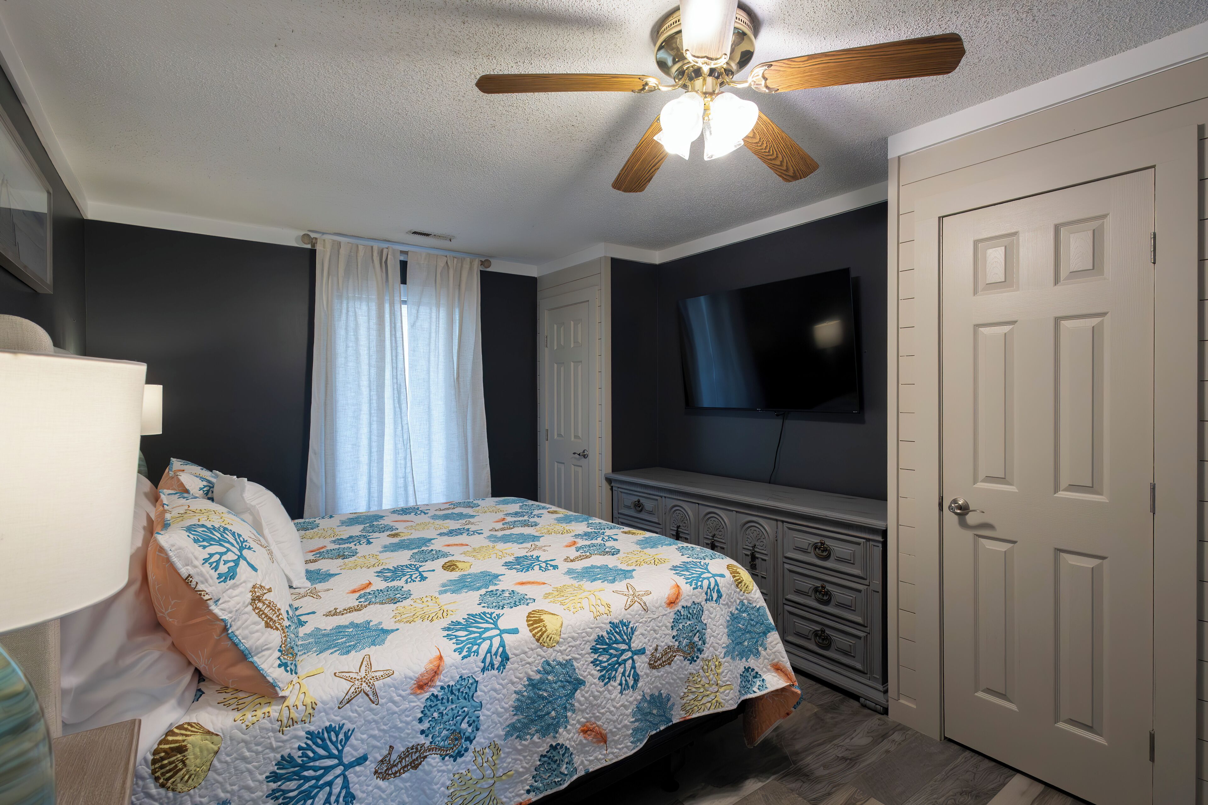 Une chambre à coucher avec un lit, un ventilateur de plafond, une commode, un téléviseur et une porte.