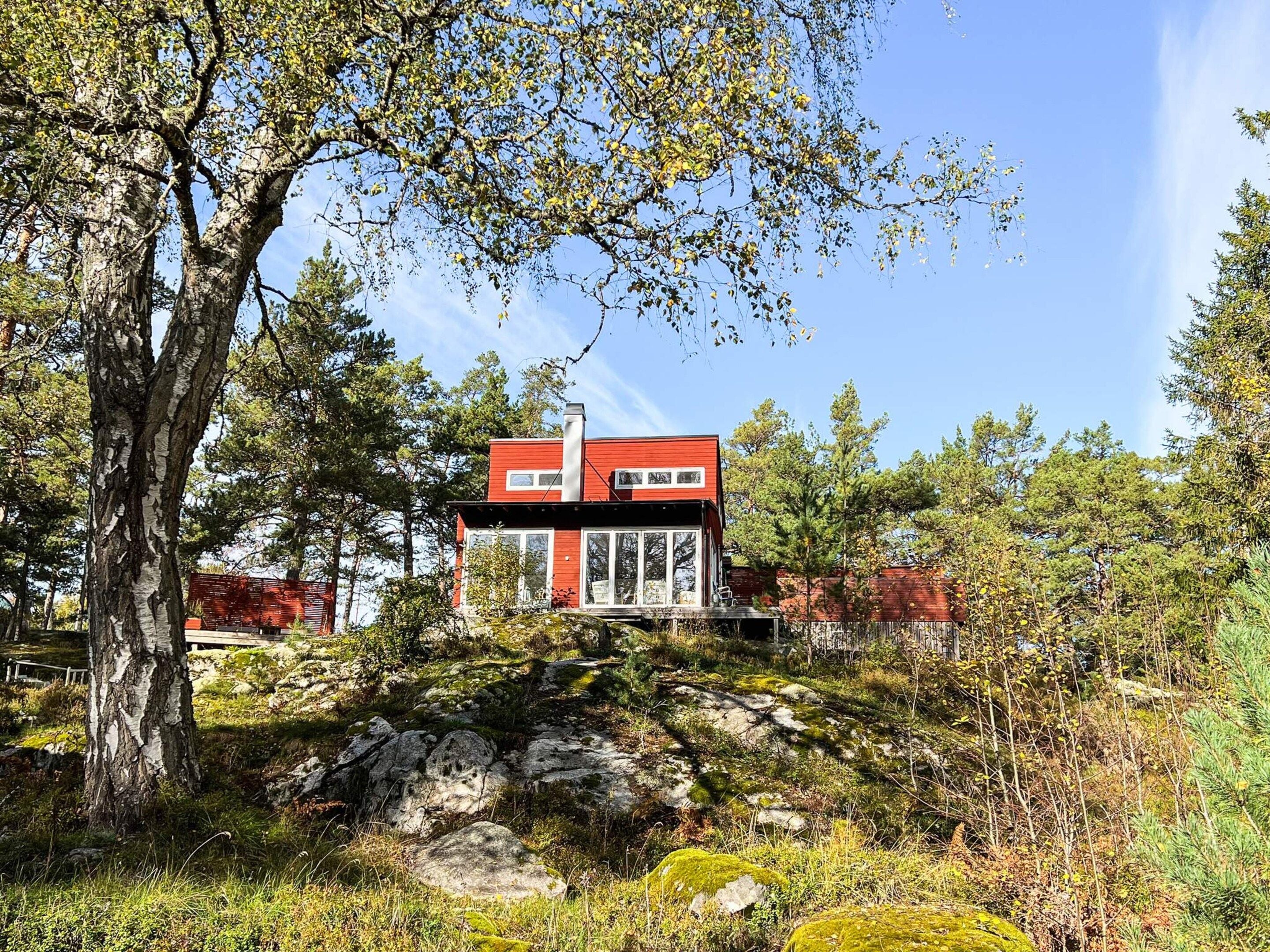Uma casa moderna com fachada vermelha, localizada em terreno rochoso rodeado por árvores.