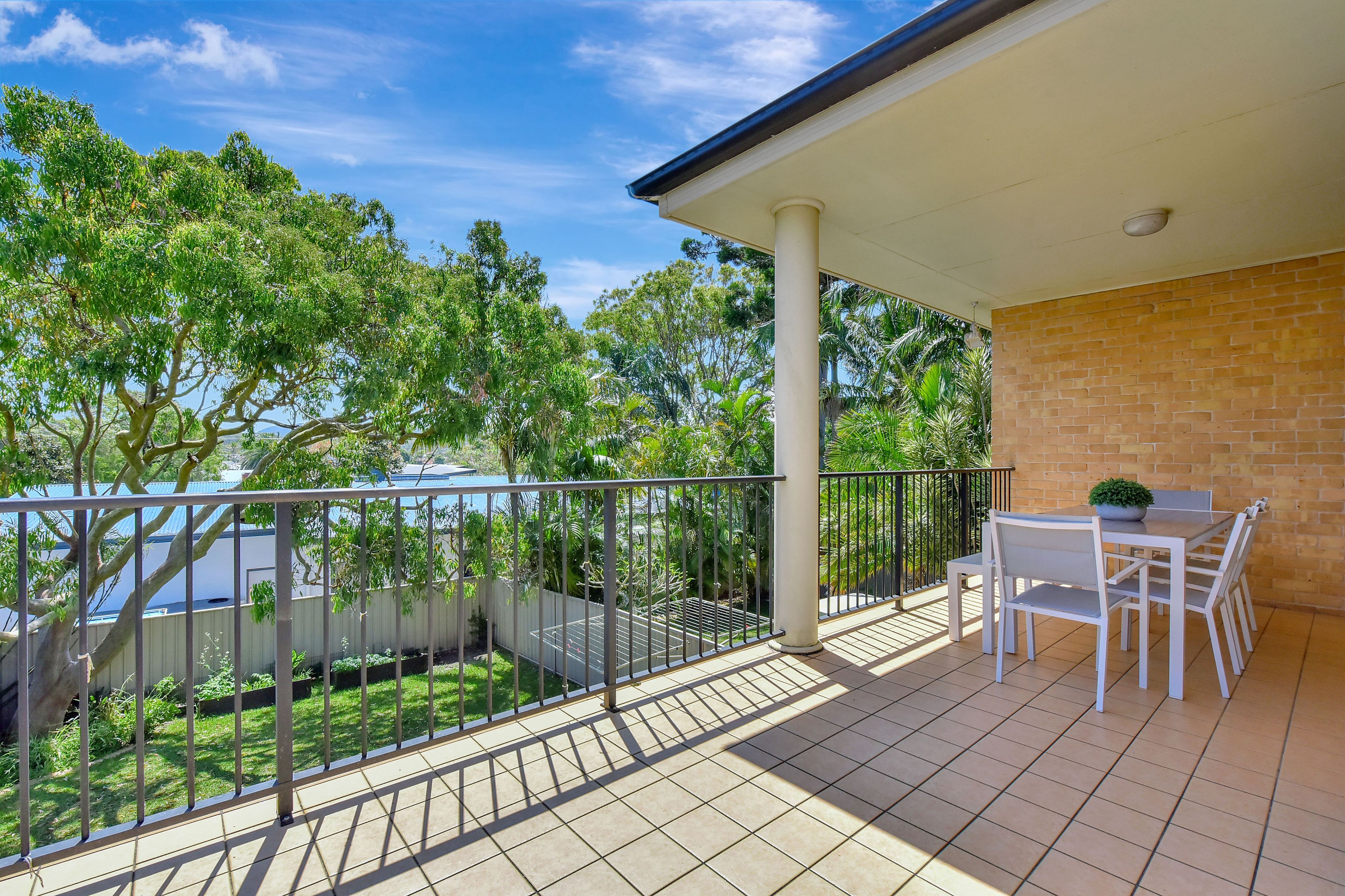 A covered outdoor area with a tiled floor, a table with chairs, and a view of greenery and a building in the background.