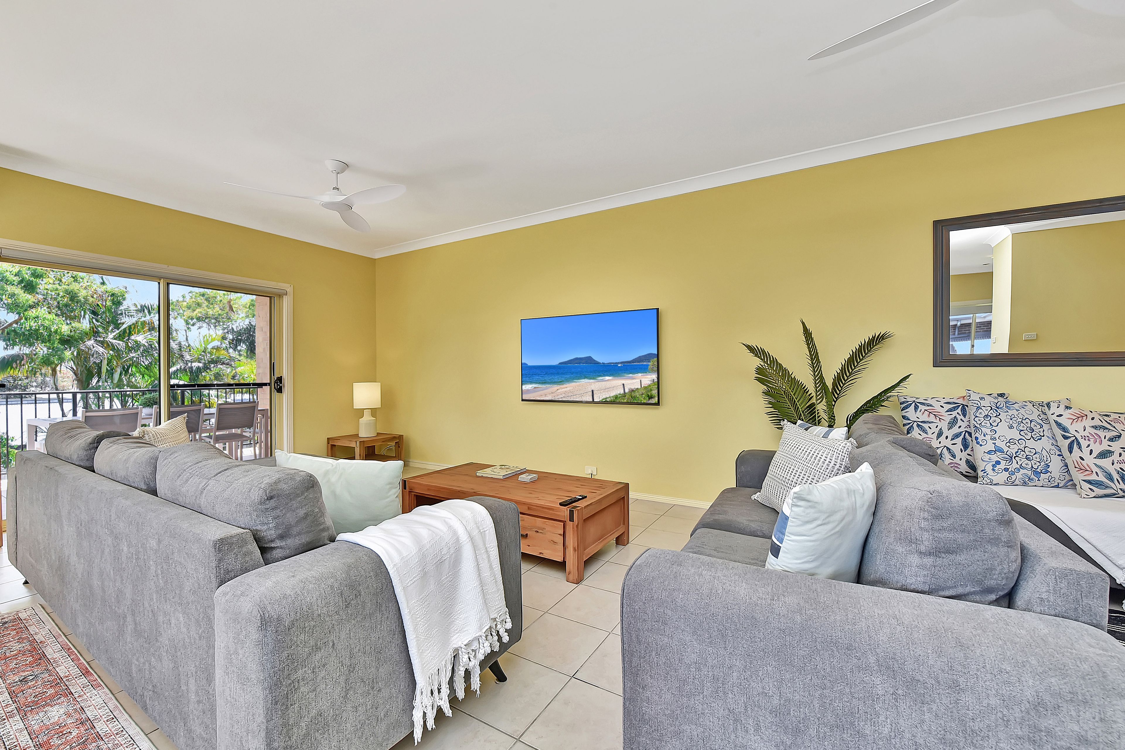 A living room with two grey sofas, a wooden coffee table, a TV on the wall, and a balcony with a view of greenery.