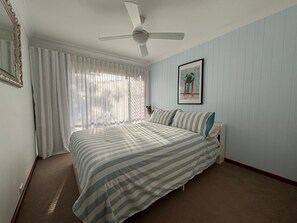 Ein Schlafzimmer mit einem Bett, einem Deckenventilator, einem Fenster mit durchscheinenden Vorhängen, einem gerahmten Bild an der Wand und einem Teppichboden.