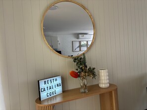 Ein Spiegel mit Holzrahmen, ein Schild mit der Aufschrift 'REST @ CATALINA COVE', eine Vase mit Blumen und ein gestreifter Kerzenständer auf einer Holzkonsole.