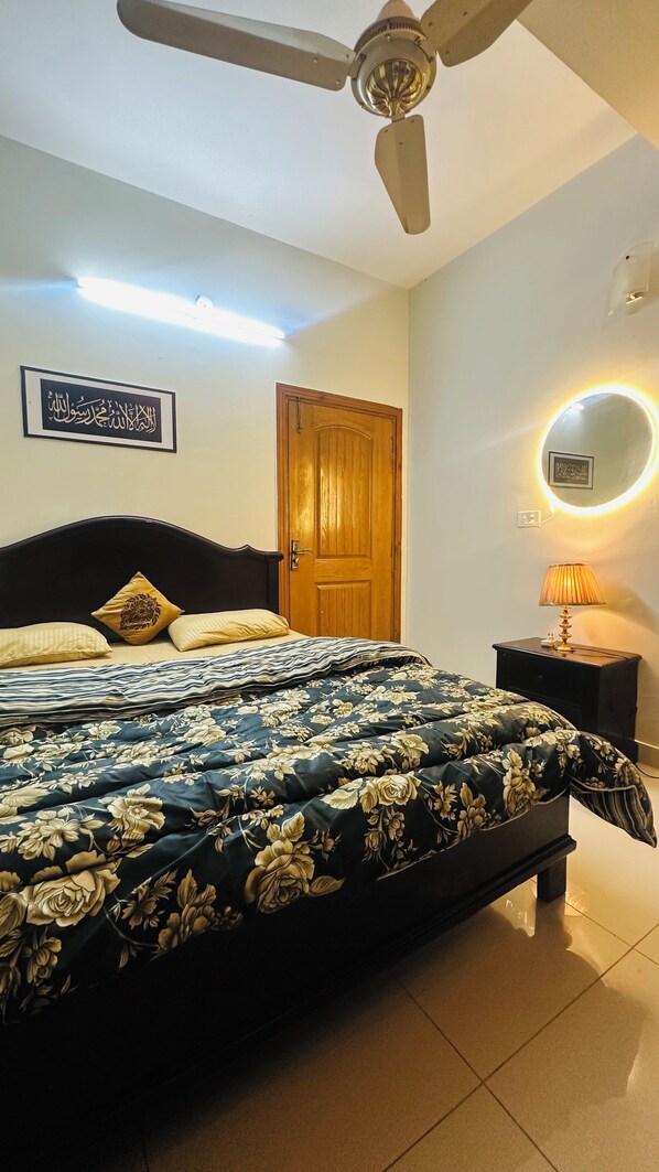 A bedroom with a bed, a ceiling fan, a wooden door, a bedside table with a lamp, and a wall-mounted light fixture.