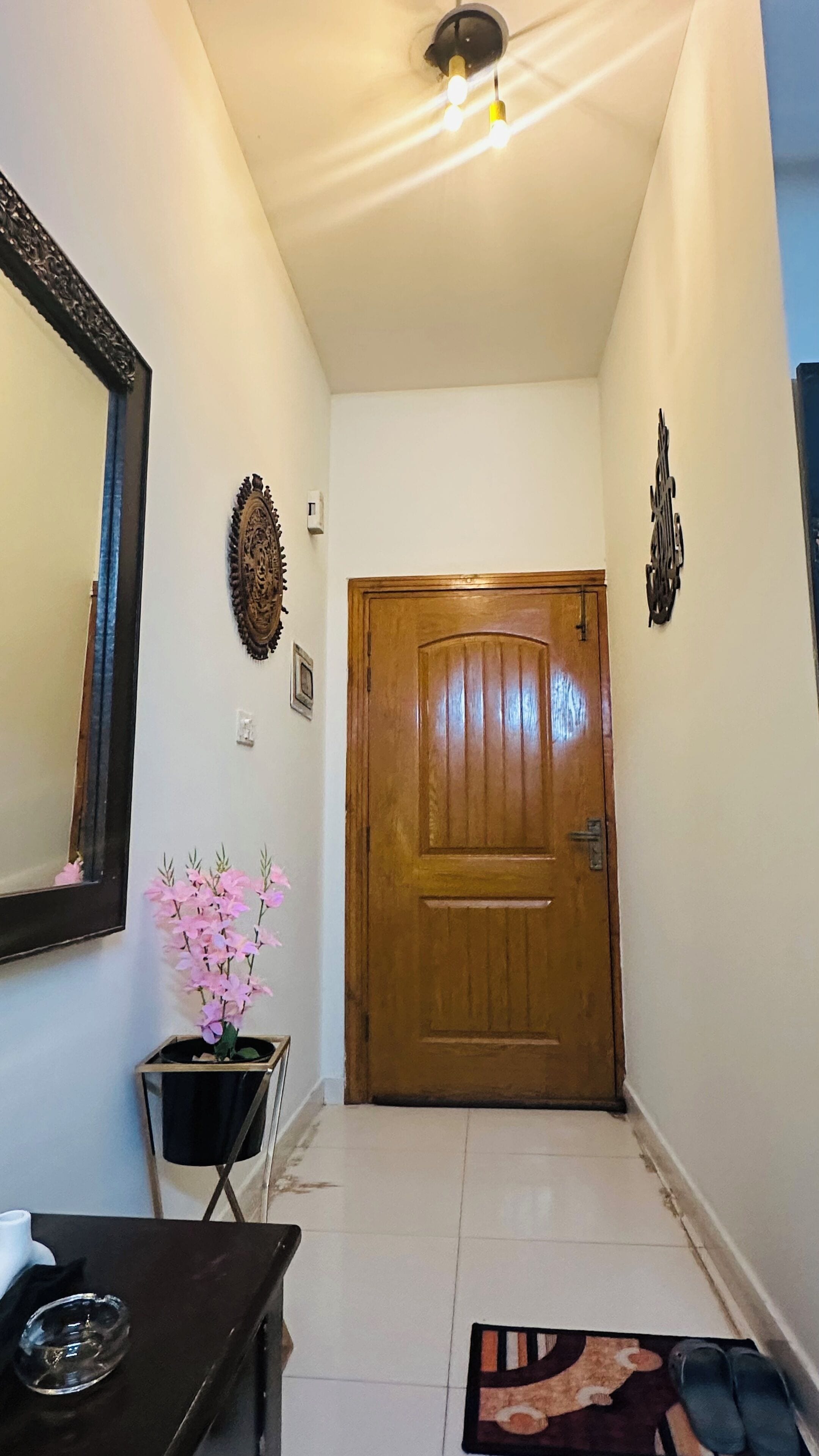 Un couloir avec une porte en bois, un miroir, une orchidée en pot et une petite table sur laquelle se trouve un cendrier en verre.