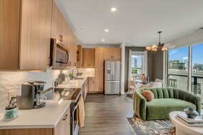 A modern kitchen with a built-in microwave, stainless steel refrigerator, and a coffee machine. There is a dining area with a green sofa and a table, and a balcony with a view of the city. - Cozy Stay ATL -2BD/2BTH FreeParking & 5* Amenities (Atlanta)
