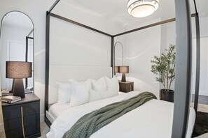 Ein Bett mit Baldachin, belegt mit weißen Leinentüchern, zwei Nachttischlampen, ein dunkler Nachttisch, ein Spiegel und ein Topfpflanze.