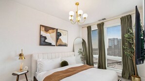 Un dormitorio con una cama grande, una mesita de noche con lámpara, un espejo y dos cuadros abstractos en la pared.