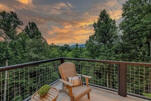 A deck with a wicker chair and a small table, surrounded by a metal railing, overlooking a forested landscape.